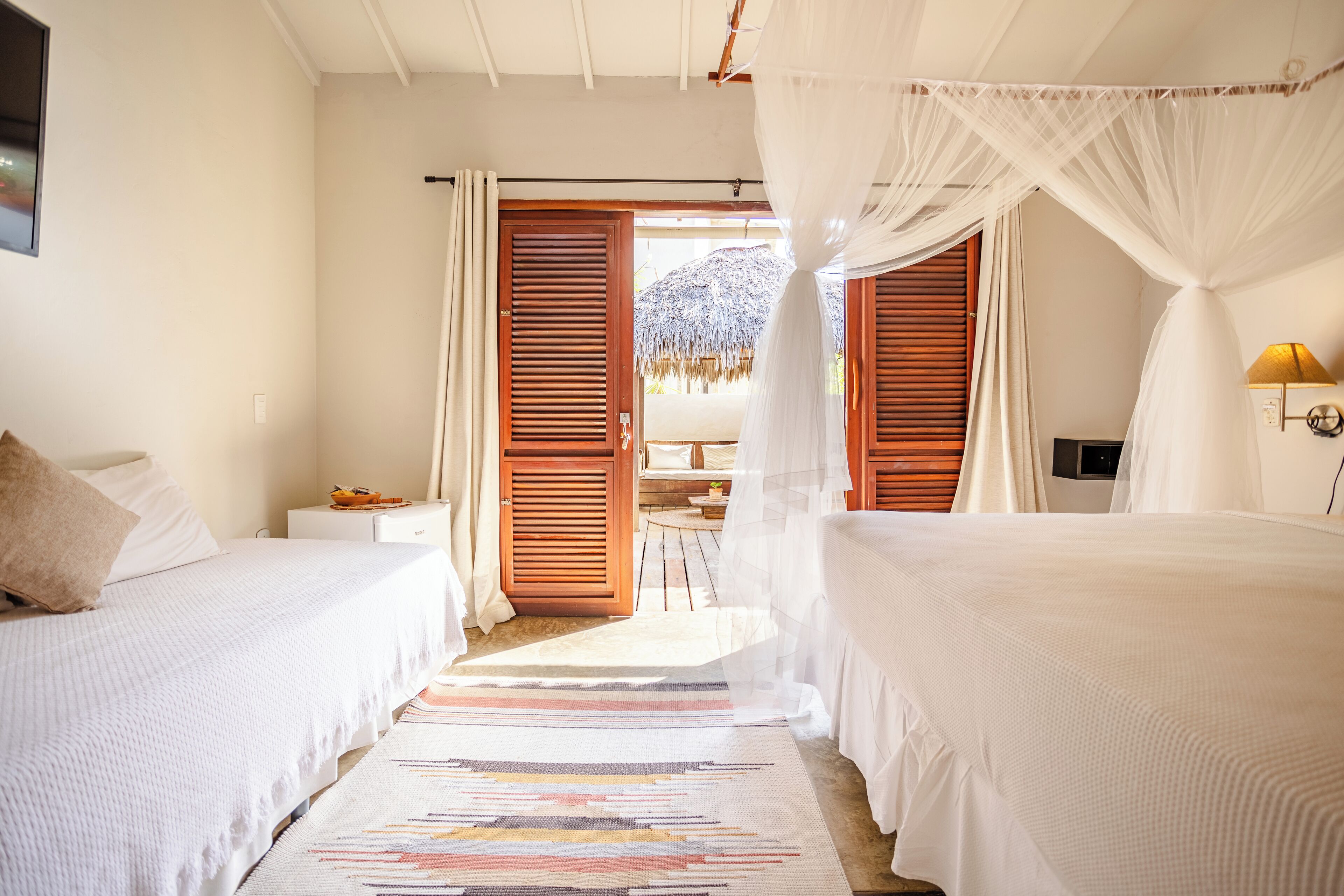 Una habitación con dos camas, una puerta de madera que da al exterior, una mesita con una lámpara y vista a una construcción de paja.