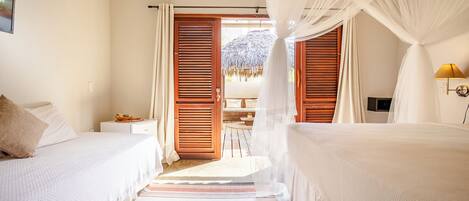 Una habitación con dos camas, una puerta de madera que da al exterior, una mesita con una lámpara y vista a una construcción de paja.
