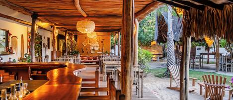 Un área de bar con barra de madera y diversas opciones de asientos, vista del patio exterior y ambiente tropical con plantas y una hamaca.
