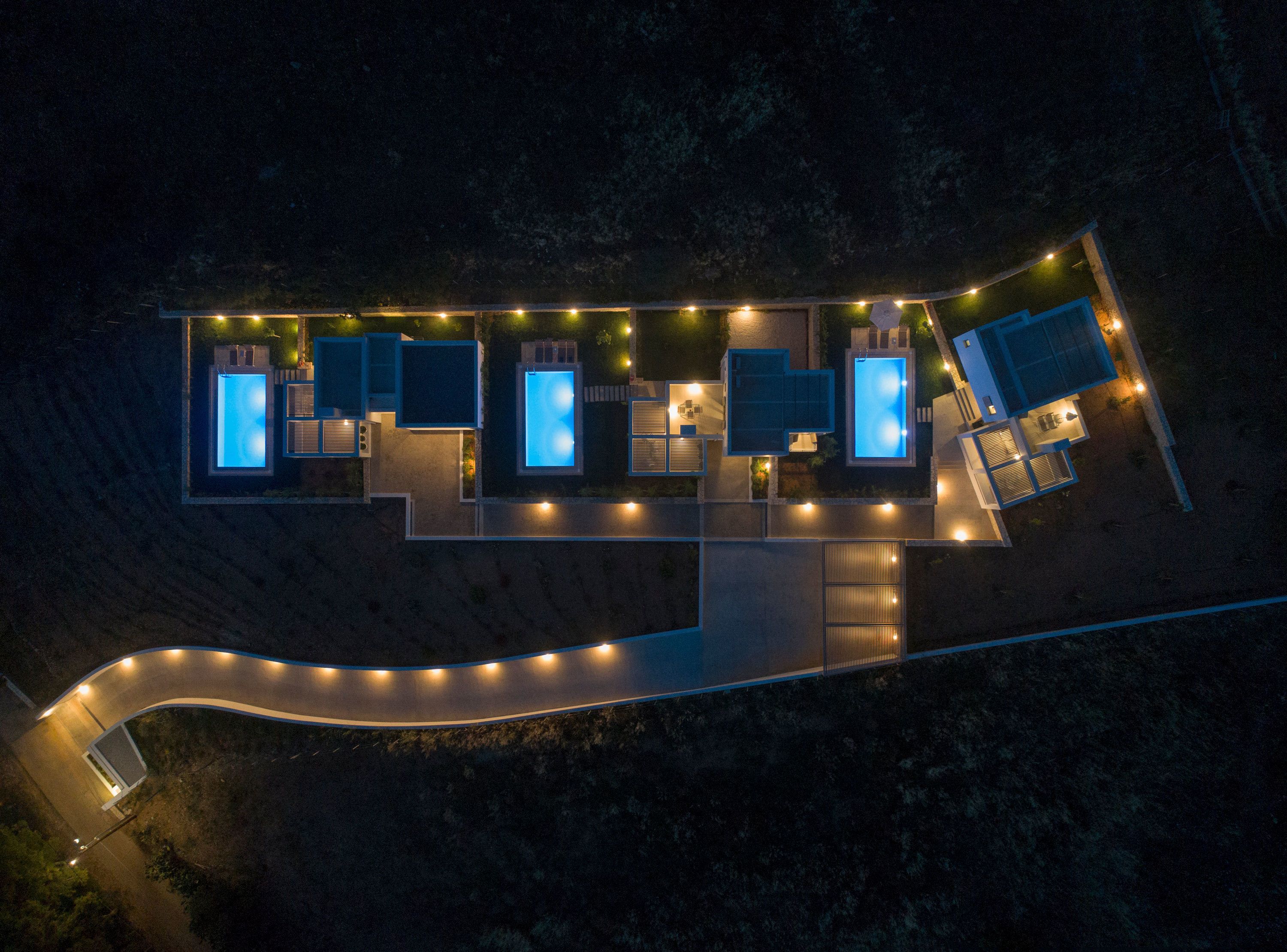An aerial view of a modern house with multiple swimming pools, a curved driveway, and outdoor lighting.