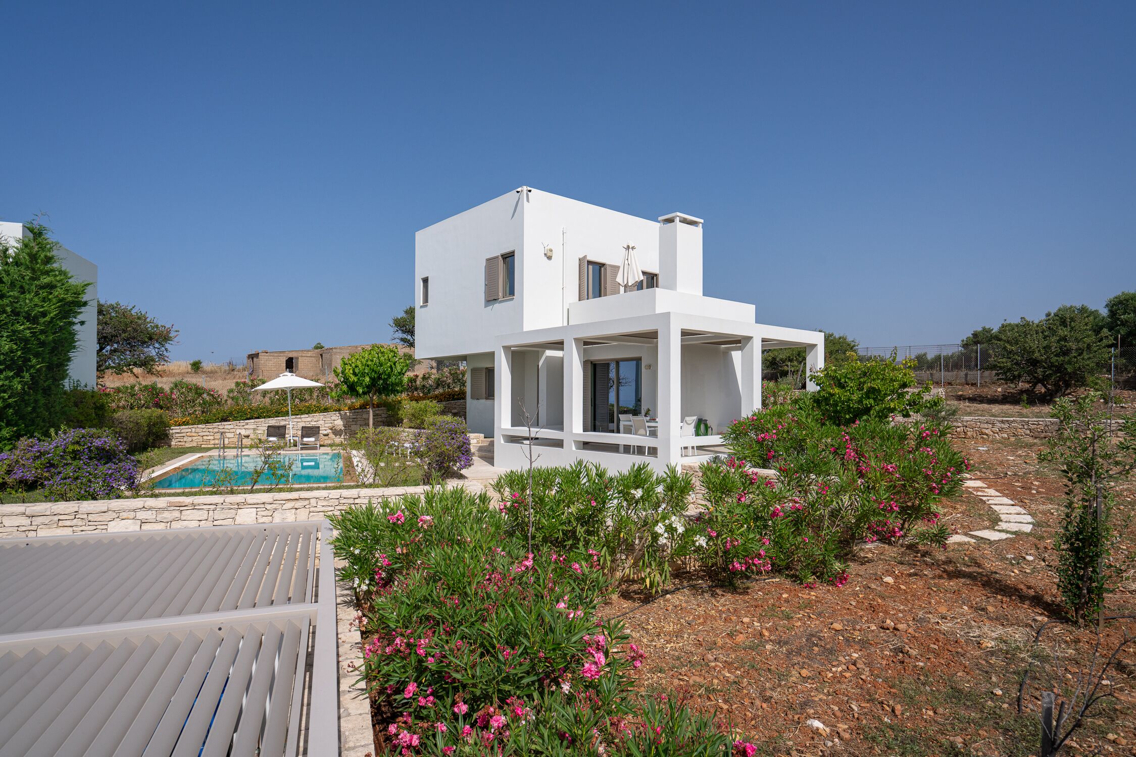 Une villa moderne aux murs blancs, dotée d’une piscine, entourée de jardins aménagés et surplombée d’un ciel d’un bleu limpide.
