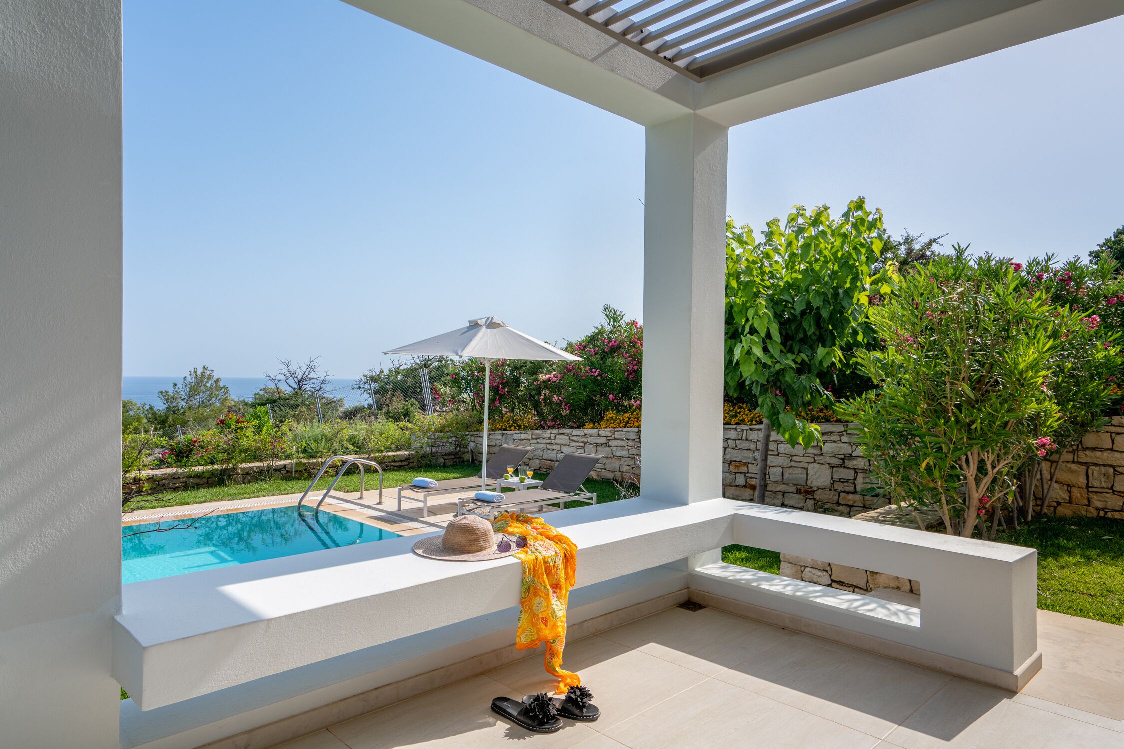 A poolside area with a view of the sea, a white pergola, a lounge chair, and a table with a hat and shoes.