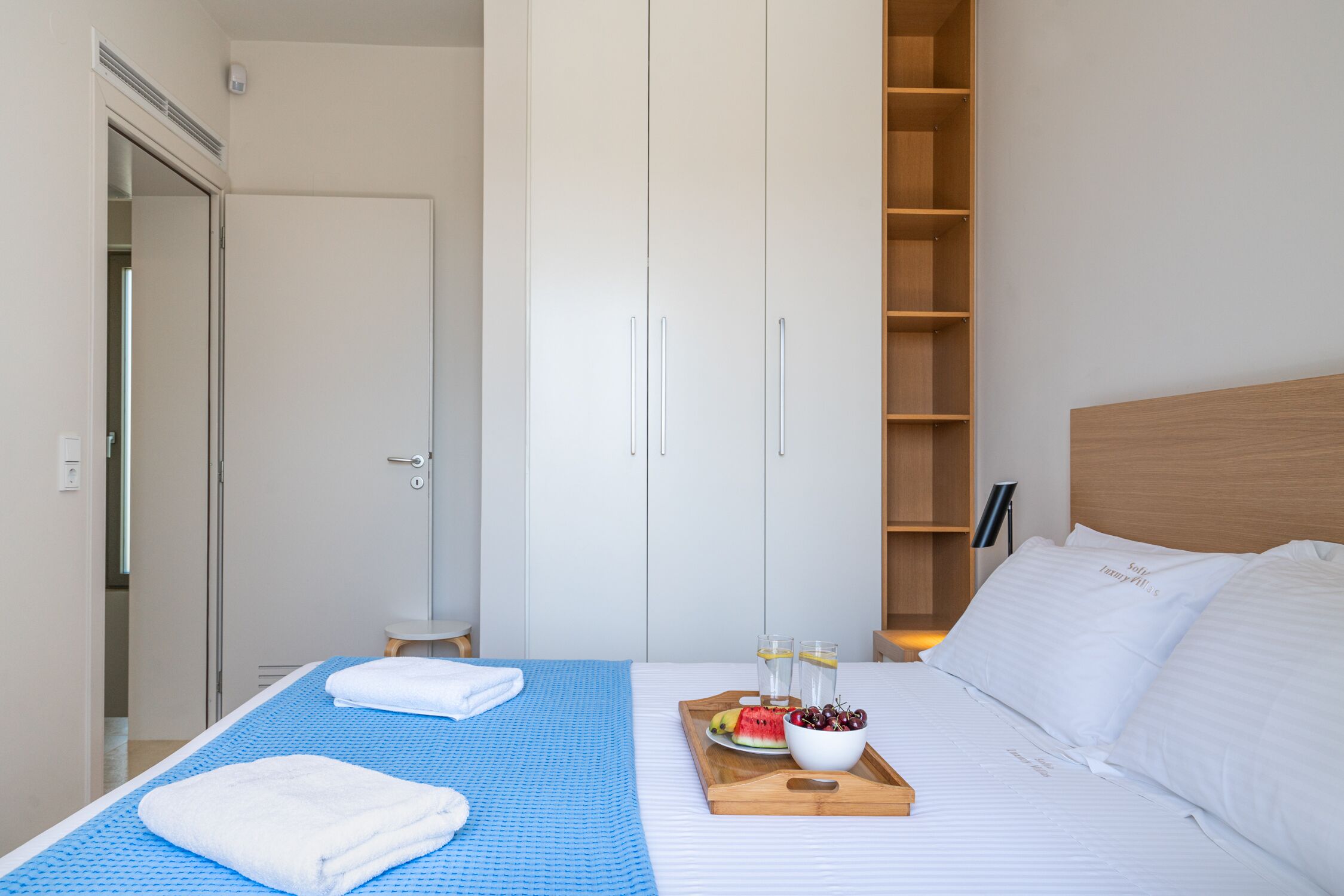 A hotel room with a bed, a wooden side table with fruit and glasses, a blue mat, and a wardrobe.