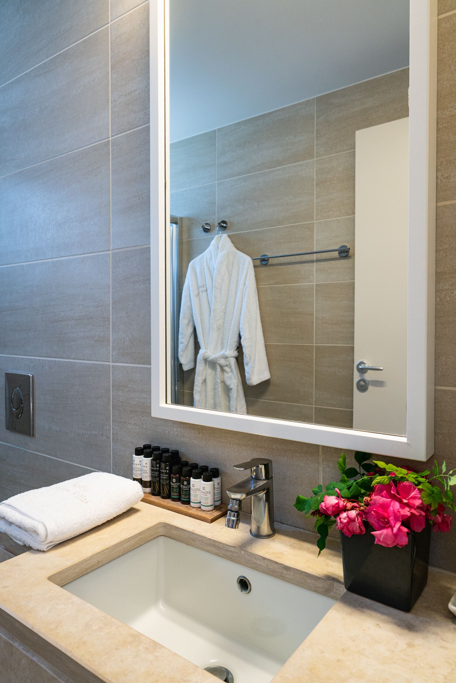 A bathroom with a large mirror, a sink, a towel, a rack of toiletries, and a potted plant.