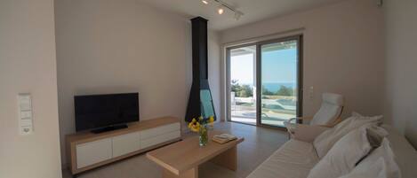 A modern living room with a flat-screen TV, a wooden coffee table, a sofa, and a large sliding glass door leading to a balcony with a view of the sea.