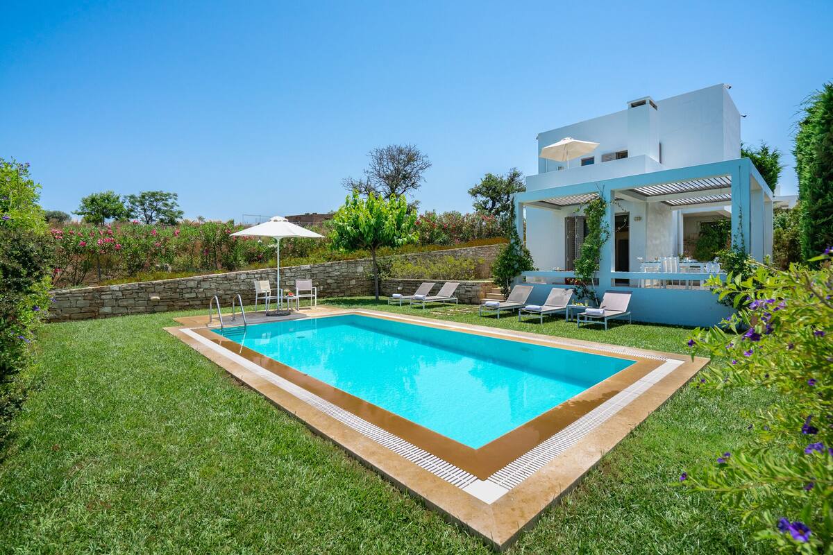 A modern pool area with a rectangular pool, surrounded by a well-maintained lawn, and a white building with a blue roof in the background.