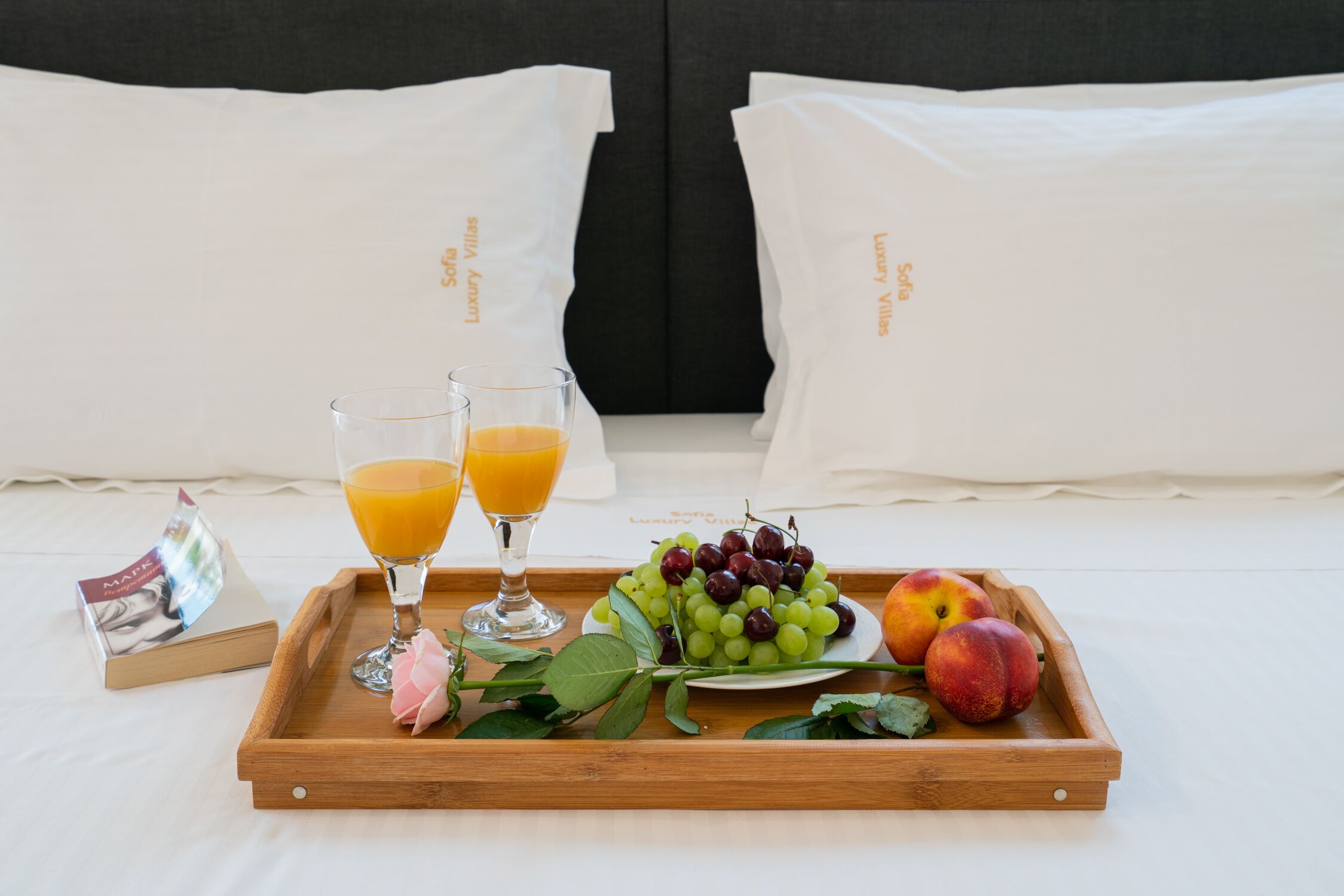 A breakfast tray with fruit and juice on a bed with white pillows and a dark headboard.