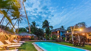 A swimming pool with lounge chairs and thatched-roof structures, surrounded by palm trees and a clear sky. - Pousada Titas (Cajueiro da Praia)