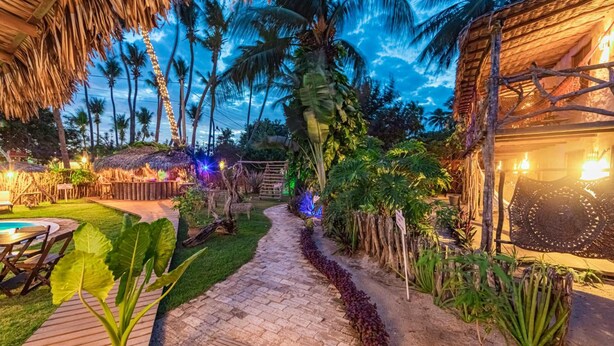 A tropical garden pathway leading to a thatched-roof structure, surrounded by lush greenery and palm trees. - Pousada Titas (Cajueiro da Praia)