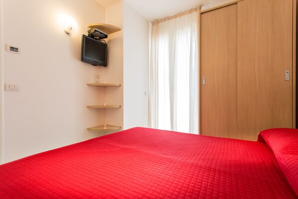 A hotel room with a red bedspread, a mounted television, and a wooden wardrobe.
