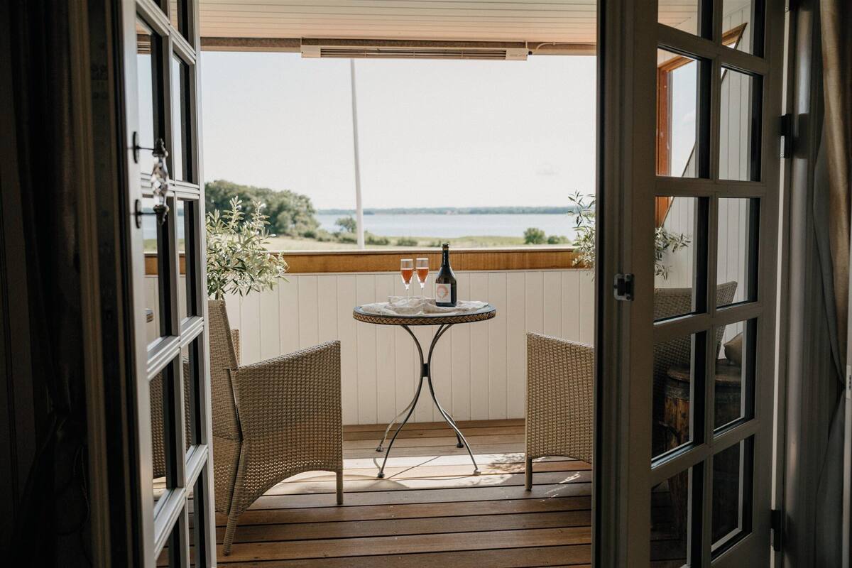 An open door leading to a balcony with wicker chairs, a small round table, and a bottle and glasses on the table.