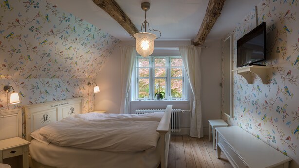 A bedroom with a bed, bedside tables, a radiator, a window with curtains, a wall-mounted TV, and a wooden ceiling. - Jungshoved Præstegård (Praesto)