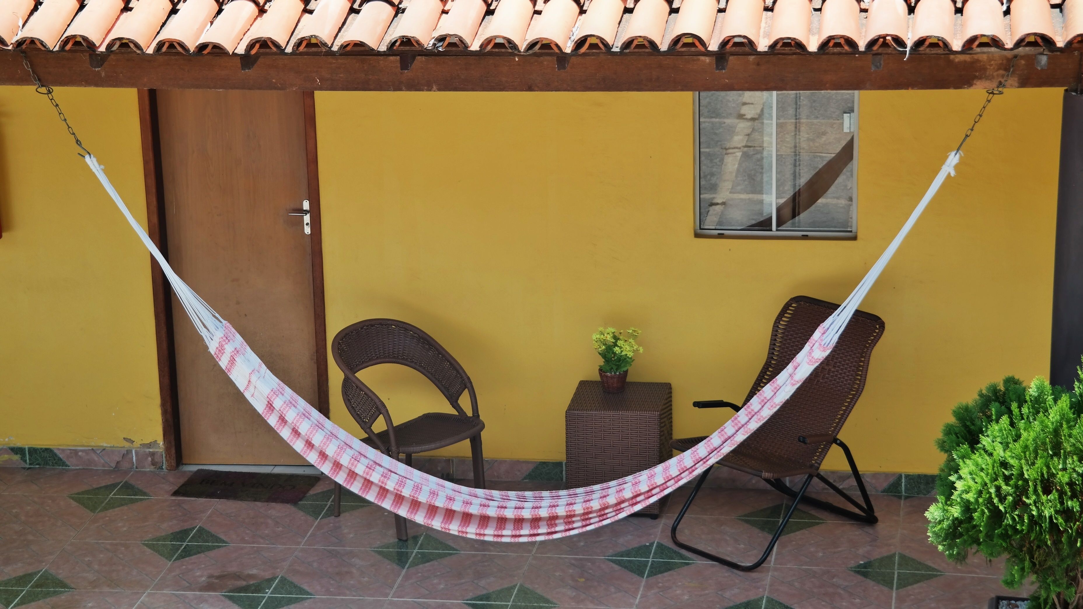 Uma rede entre duas cadeiras de vime em um pátio com piso de azulejos, uma parede amarela e uma porta.