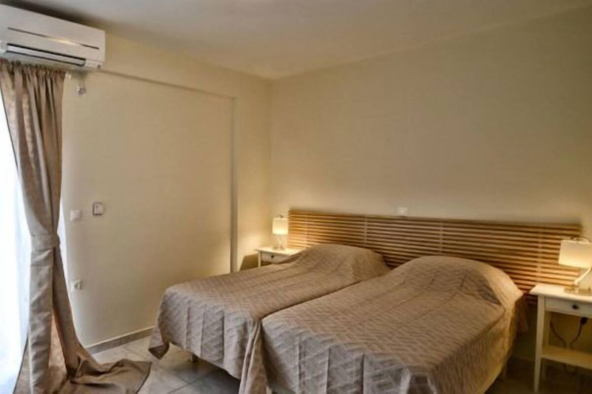 A hotel room with two beds, an air conditioning unit, a bedside table with a lamp, and curtains on the window.