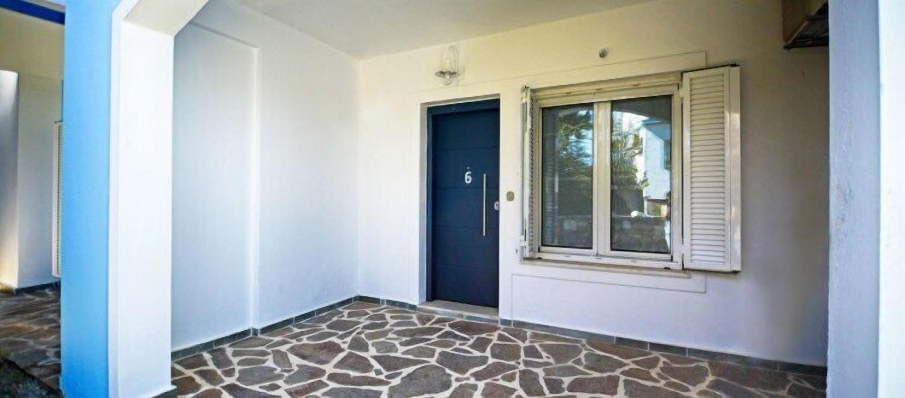 Ein Gebäudeeingang mit steinbelagtem Boden, einer dunkelblauen Tür und einem Fenster mit Fensterläden.
