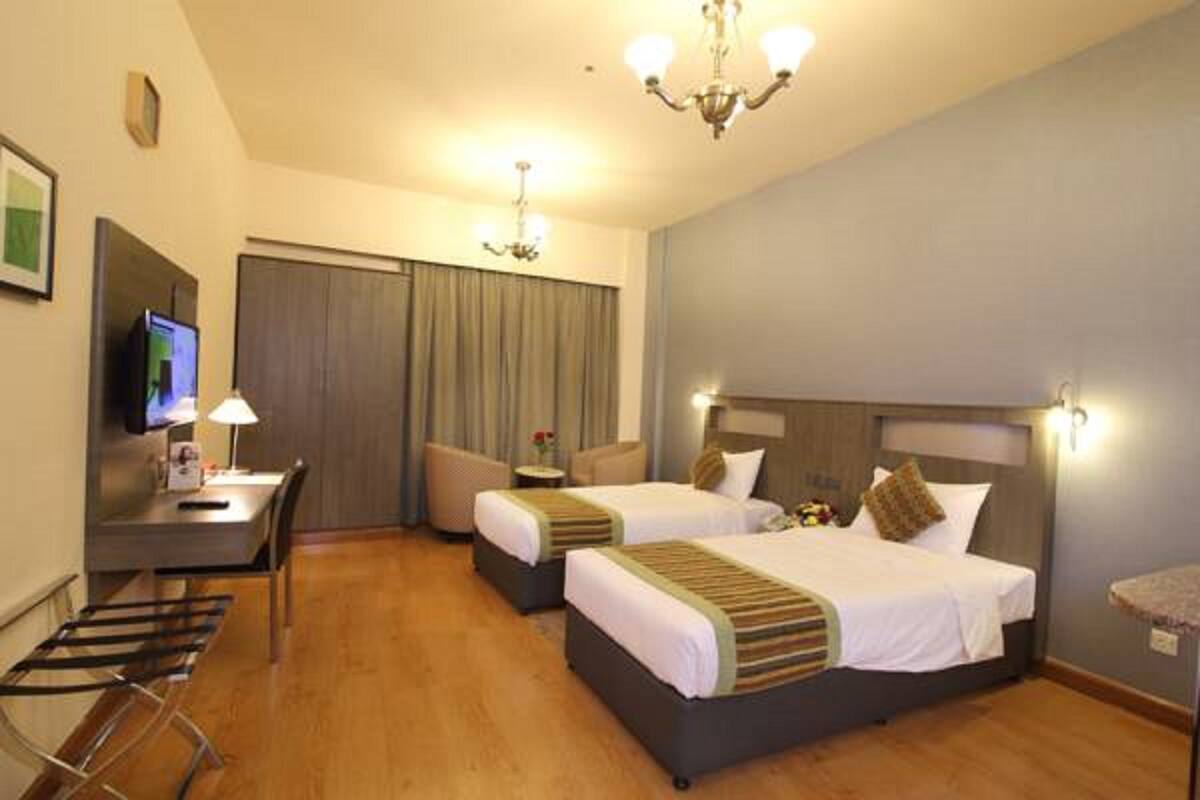 A hotel room with two beds, a desk with a computer, a chair, and a small table with a lamp.