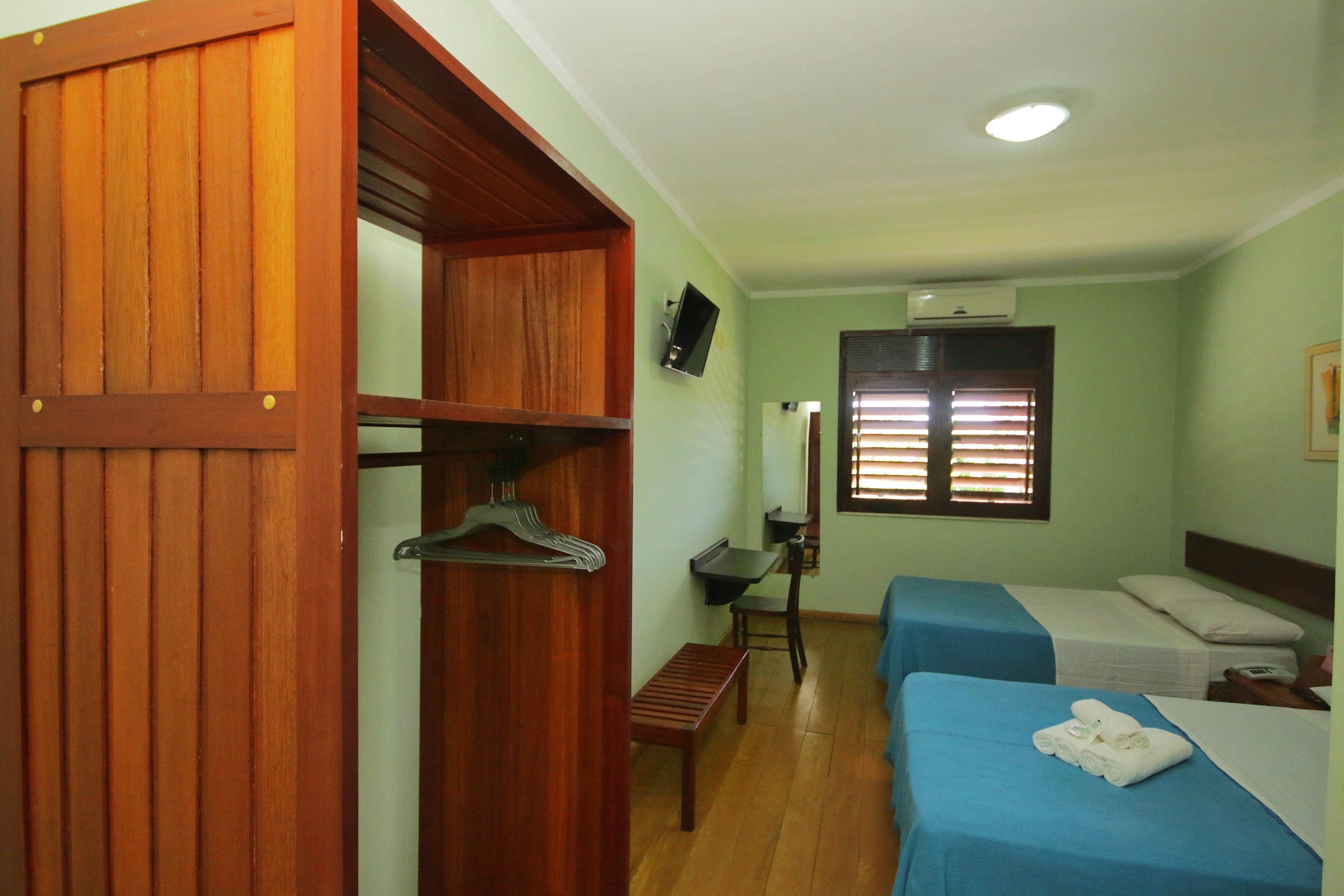 A hotel room with two beds, a wooden wardrobe, a desk with a chair, and a TV mounted on the wall.