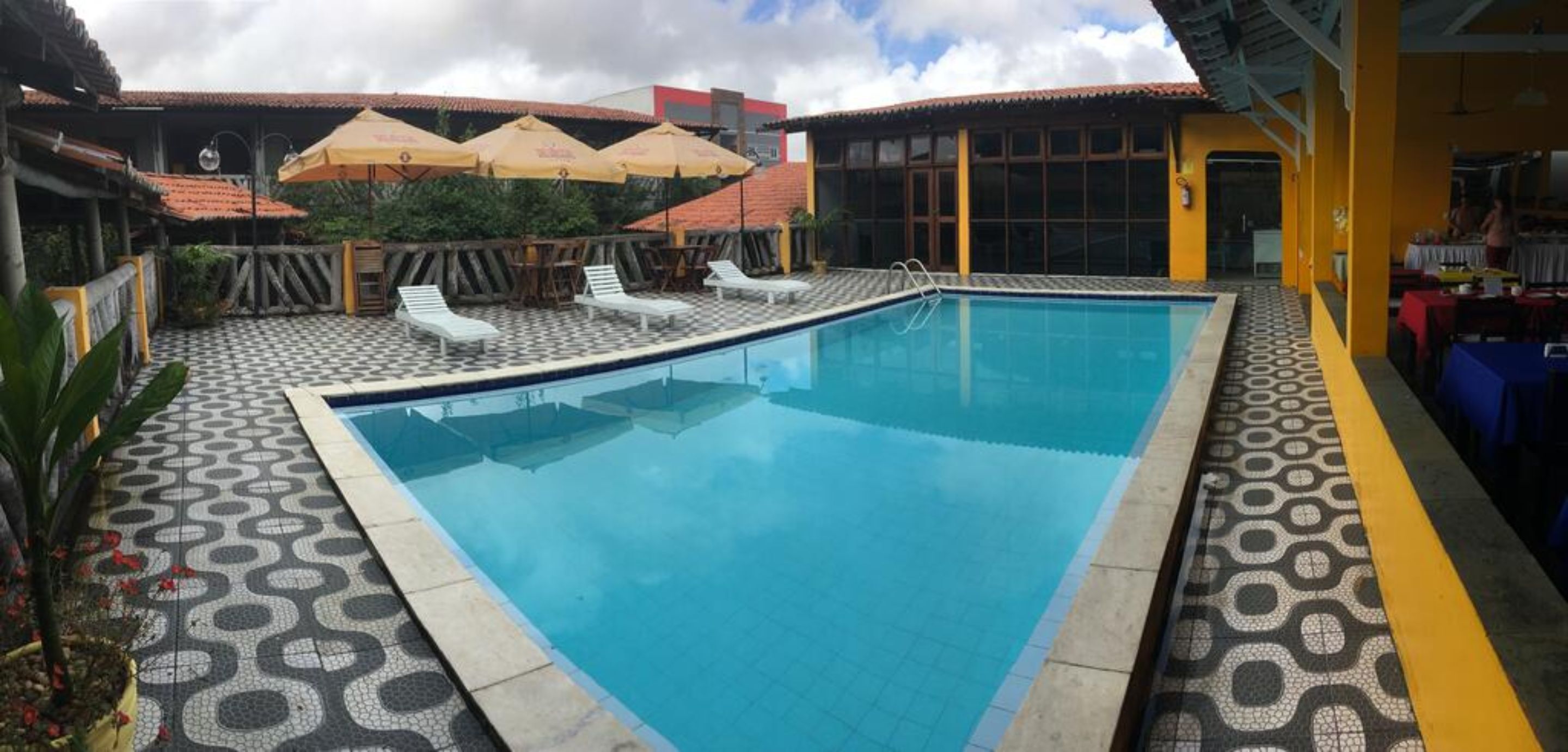 A swimming pool with lounge chairs and umbrellas, surrounded by patterned flooring and a building with a tiled roof.