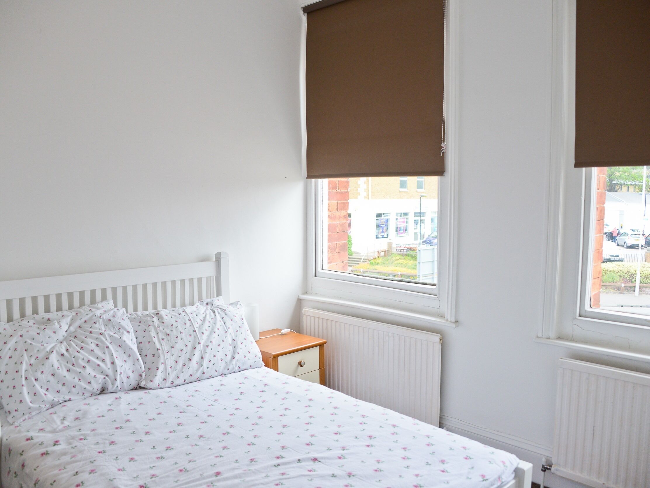 A bedroom with a bed, a nightstand, a window with a view of the street, and a radiator.