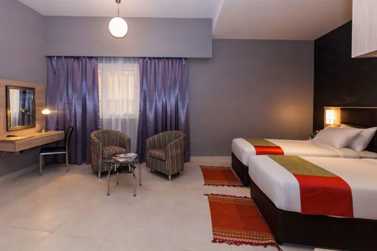 Quarto de hotel moderno com cama, duas poltronas, escrivaninha e uma janela grande com cortinas.
