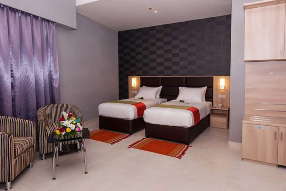 Quarto de hotel com duas camas, uma poltrona listrada, uma mesinha de centro com um vaso de flores e parede quadriculada.