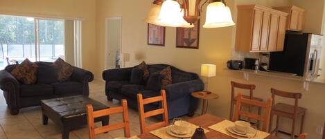 A living room with two sofas, a coffee table, and a dining table set for four. There is a kitchen area with cabinets and a refrigerator.