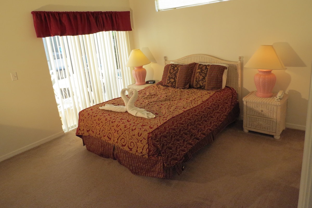 A bedroom with a large bed, two bedside lamps, a window with blinds, and a red valance.
