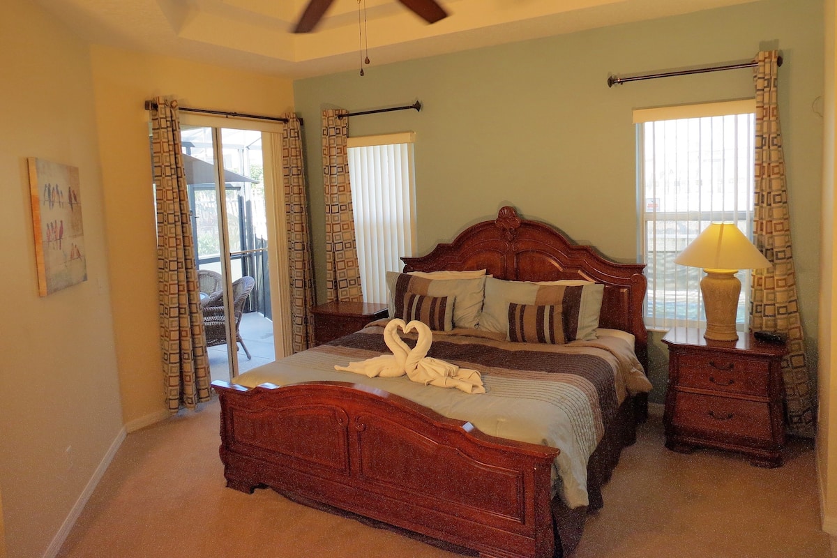A bedroom with a wooden bed, a ceiling fan, a nightstand with a lamp, and a view of the outdoors through a window.