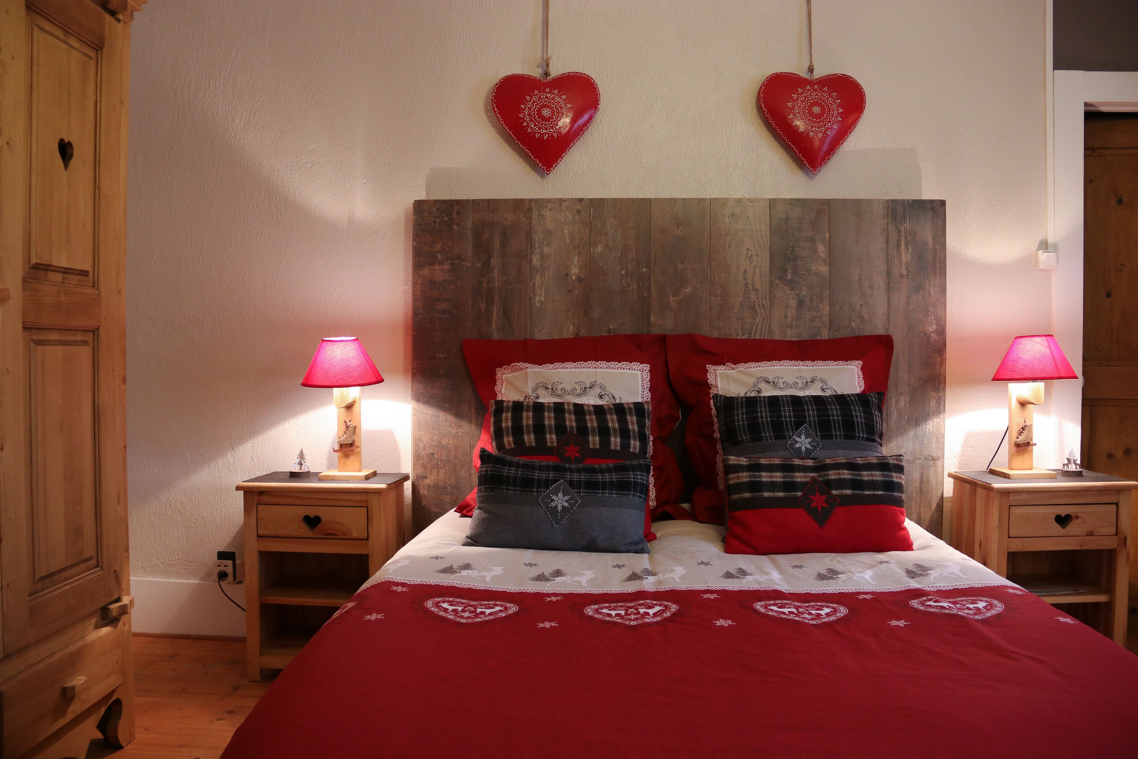 Een slaapkamer met een houten hoofdbord, twee nachtkastjes met lampen, en een bed met rode beddengoed en hartvormige kussens.