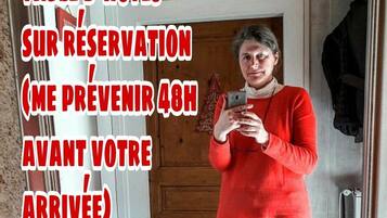 Una persona de pie en un baño, sosteniendo un teléfono, con un espejo que refleja una puerta y un taburete pequeño. Un texto superpuesto indica una reserva de hotel con un aviso de 48 horas.