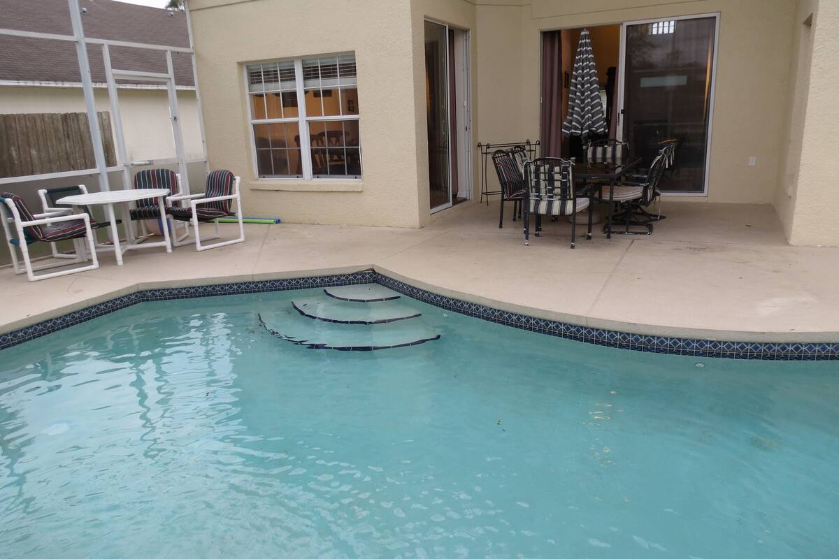 A swimming pool with steps, a patio with chairs and a table, and a sliding glass door.