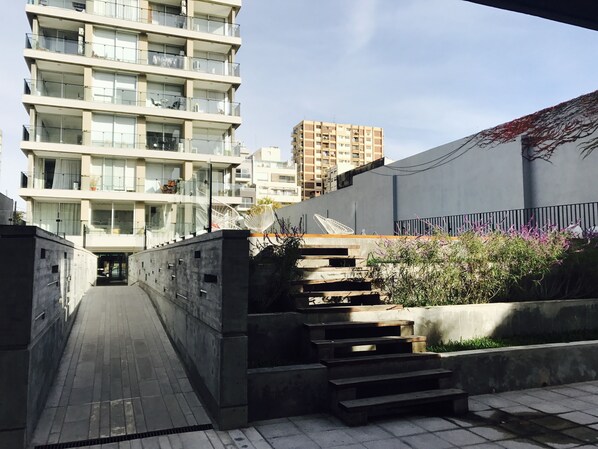 Un edificio moderno urbano con varios balcones, un estrecho pasaje y una escalera que lleva a un jardín en la azotea con plantas y una silla blanca.