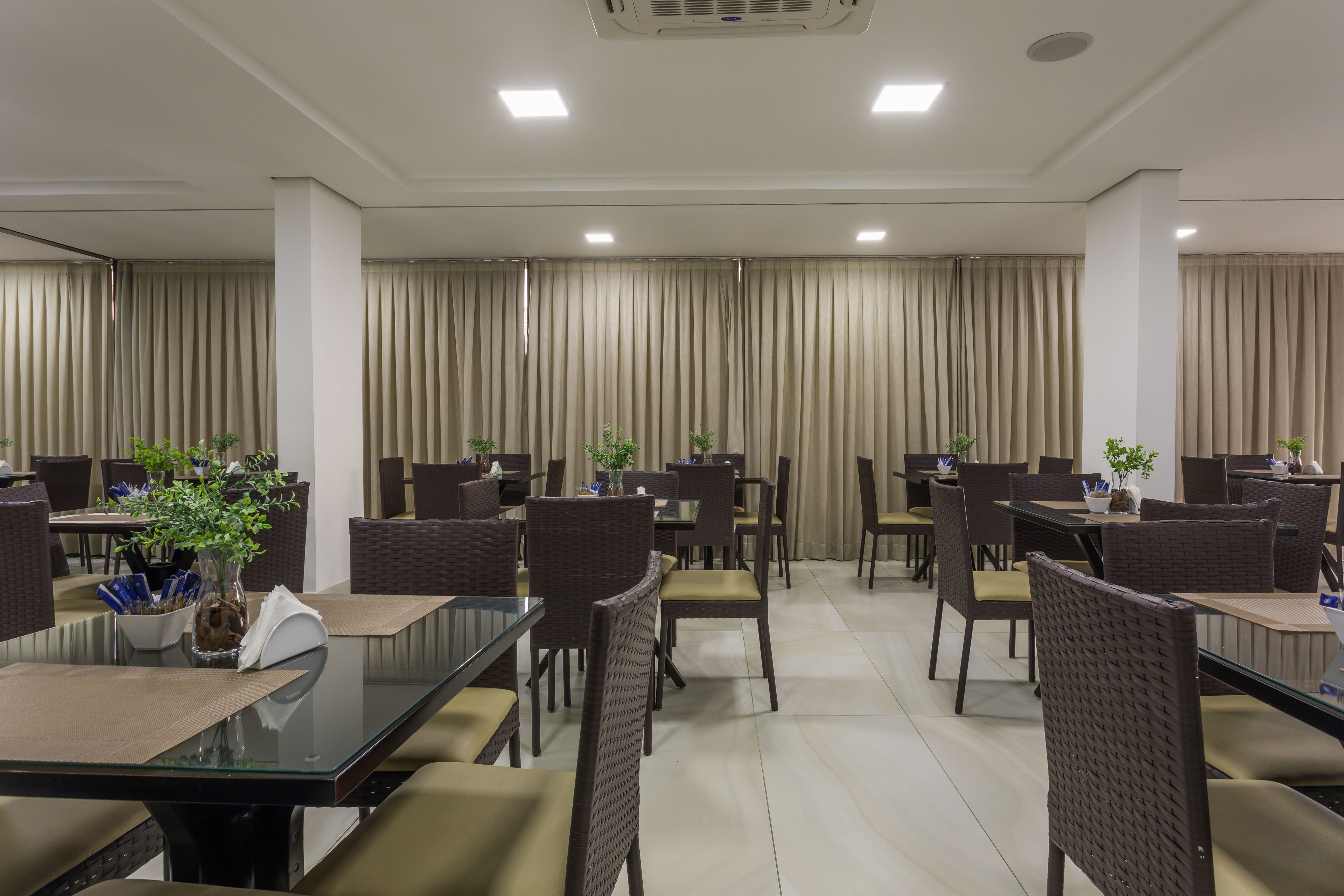 A conference room with glass tables and brown wicker chairs, beige curtains, and potted plants.