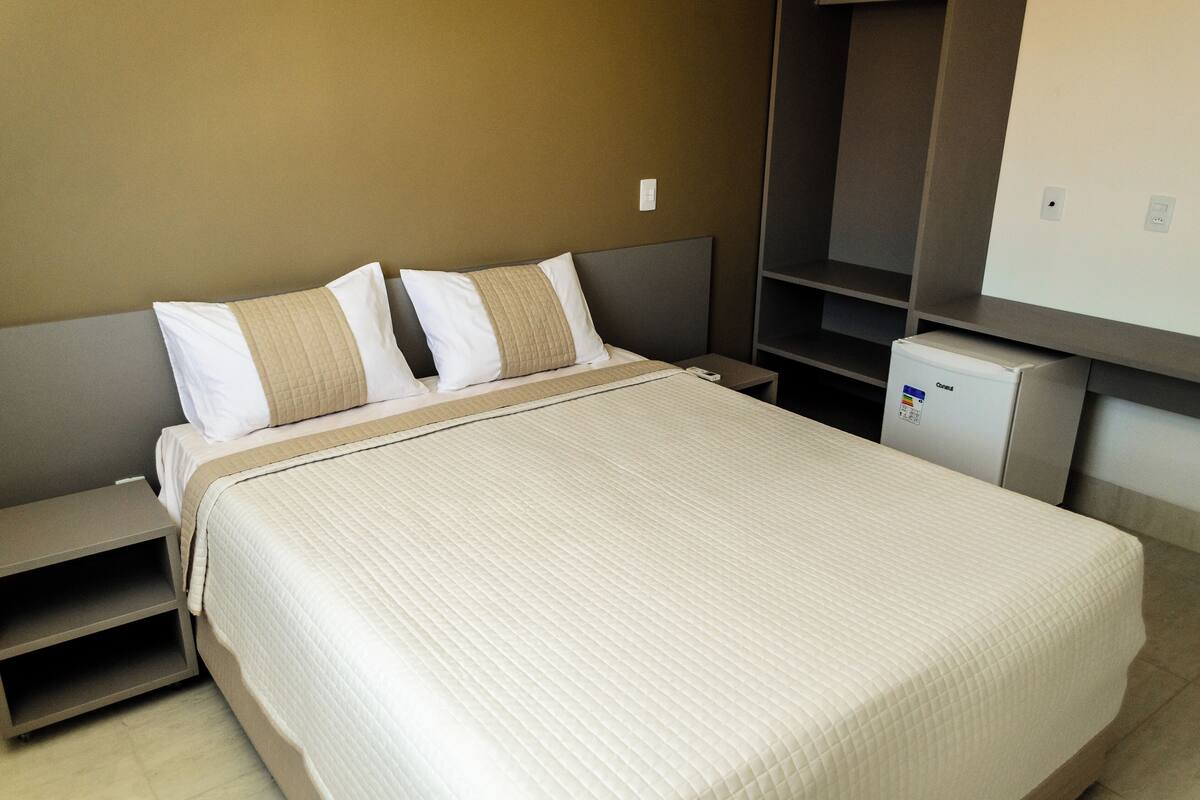 A neatly made bed with a quilted bedspread, a bedside table, and a small white appliance, likely a mini-fridge, in a hotel room with a simple, functional design.