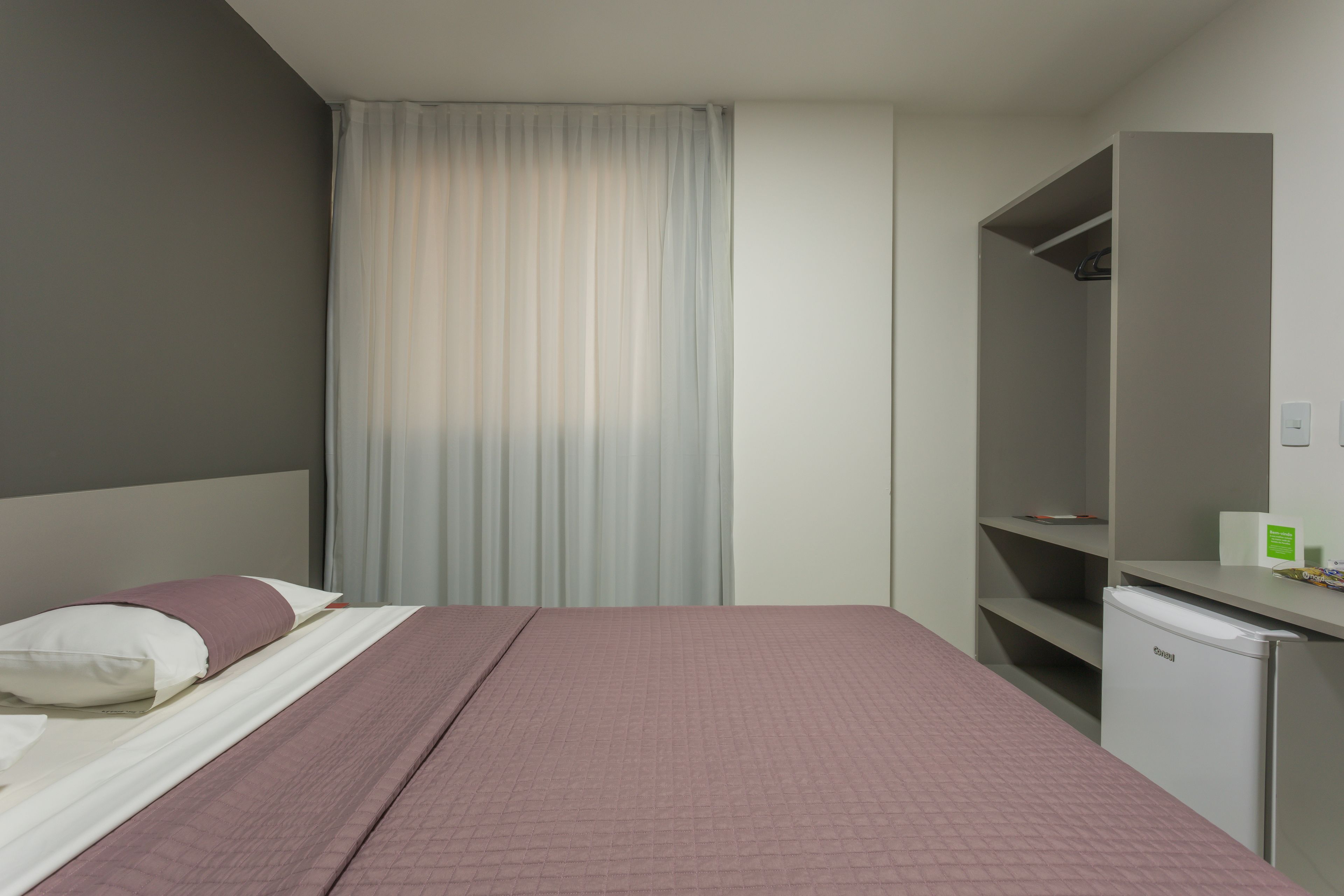 A hotel room with a bed, a desk, a mini-fridge, and a wardrobe.