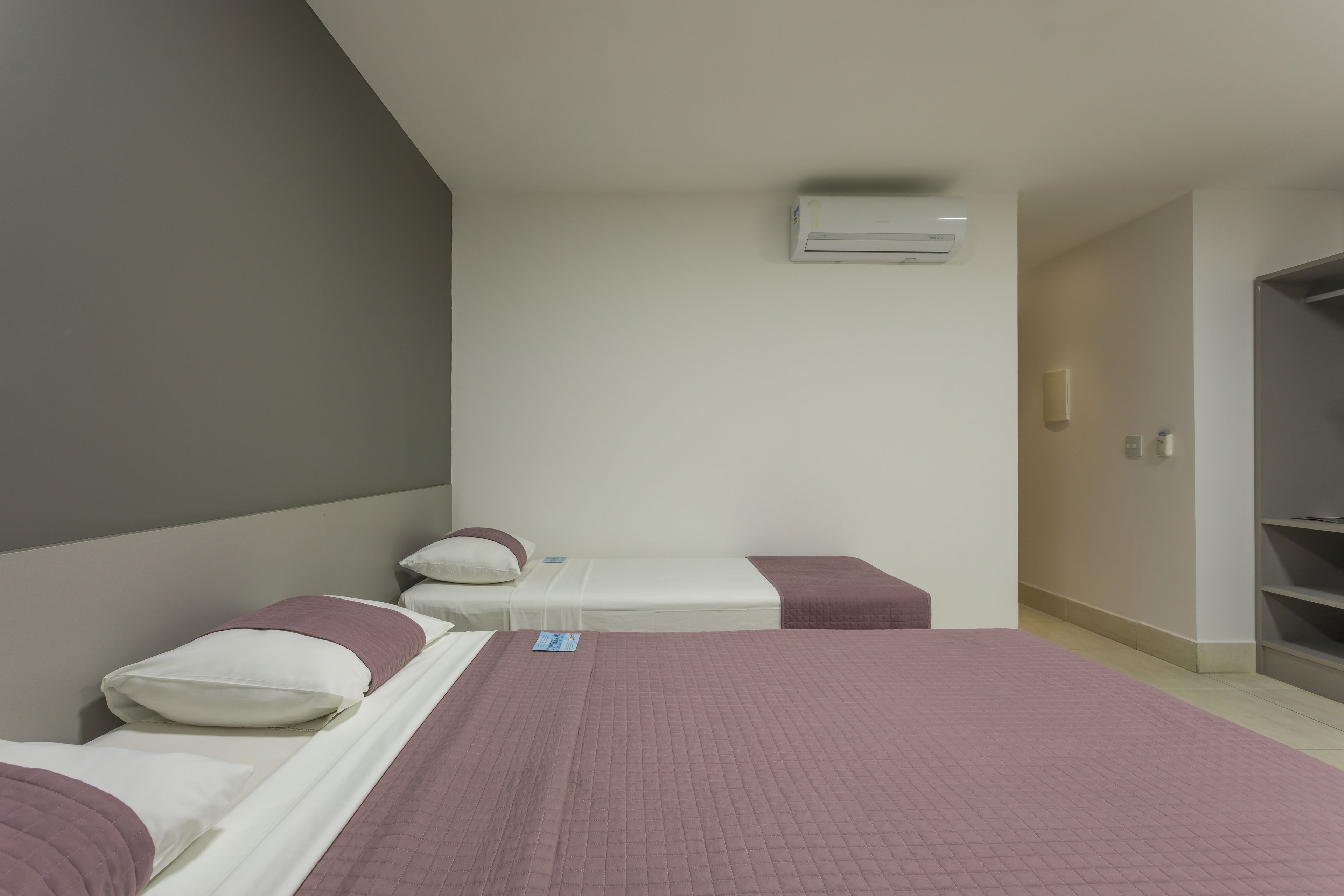 A hotel room with a large bed, two smaller beds, a wardrobe, and an air conditioning unit.