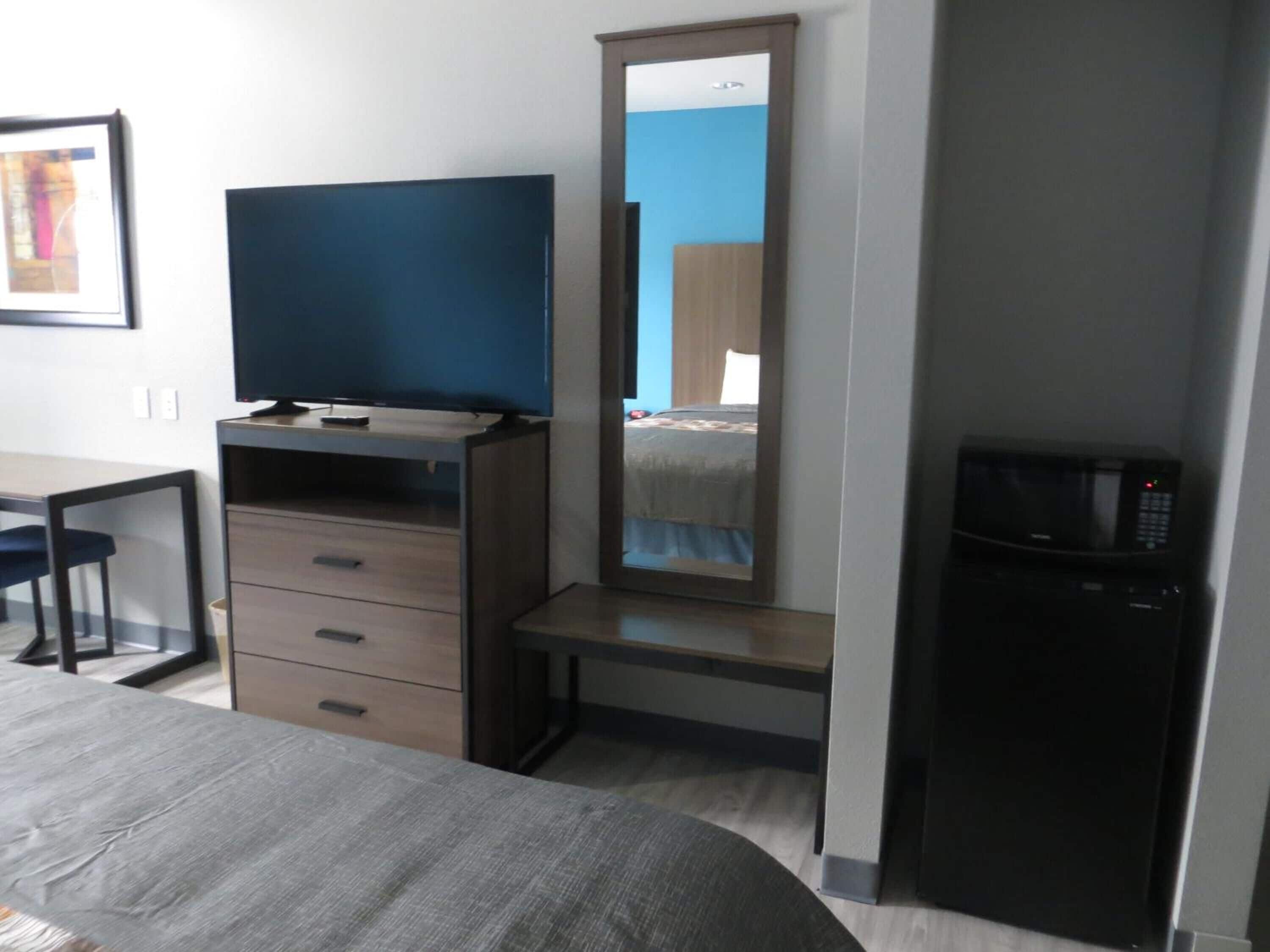 A hotel room with a bed, a desk with a chair, a dresser, a mirror, and a microwave.