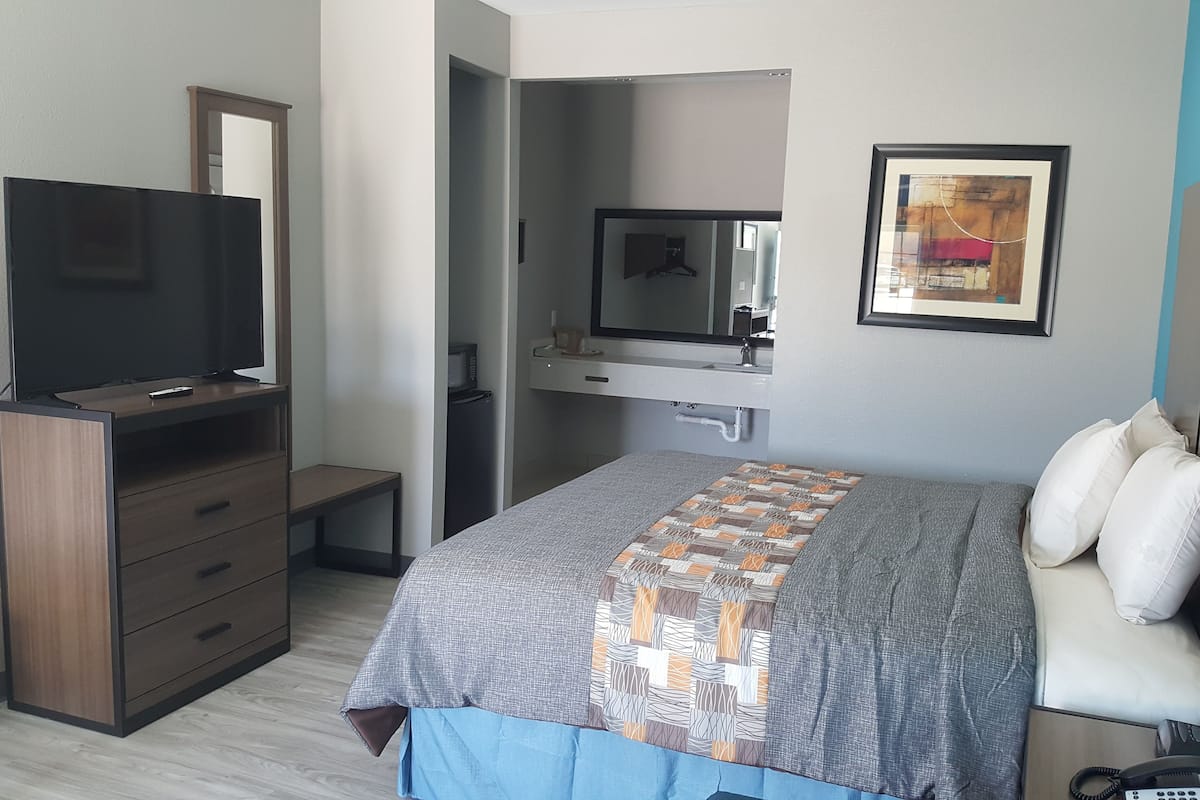 A hotel room with a bed, a TV on a wooden cabinet, a bathroom with a mirror, and a framed picture on the wall.