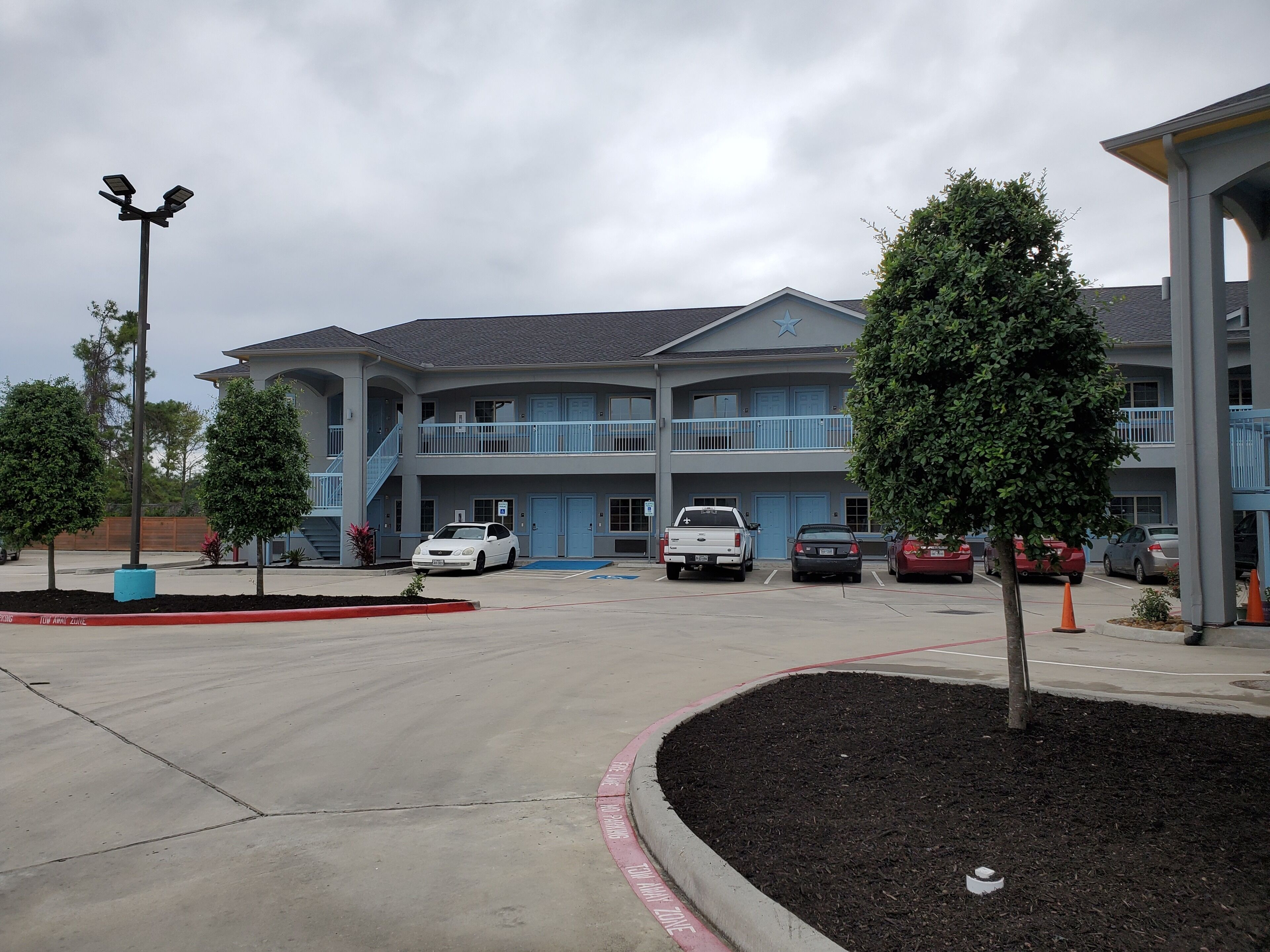 A multi-story building with a blue exterior, a parking lot with several cars, and a landscaped area with trees.