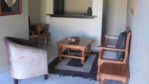 Una sala de estar con un sofá, una mesa de madera con un cuenco de fruta, una silla con cojín, un mueble de madera pequeño y una estantería de pared con un jarrón.