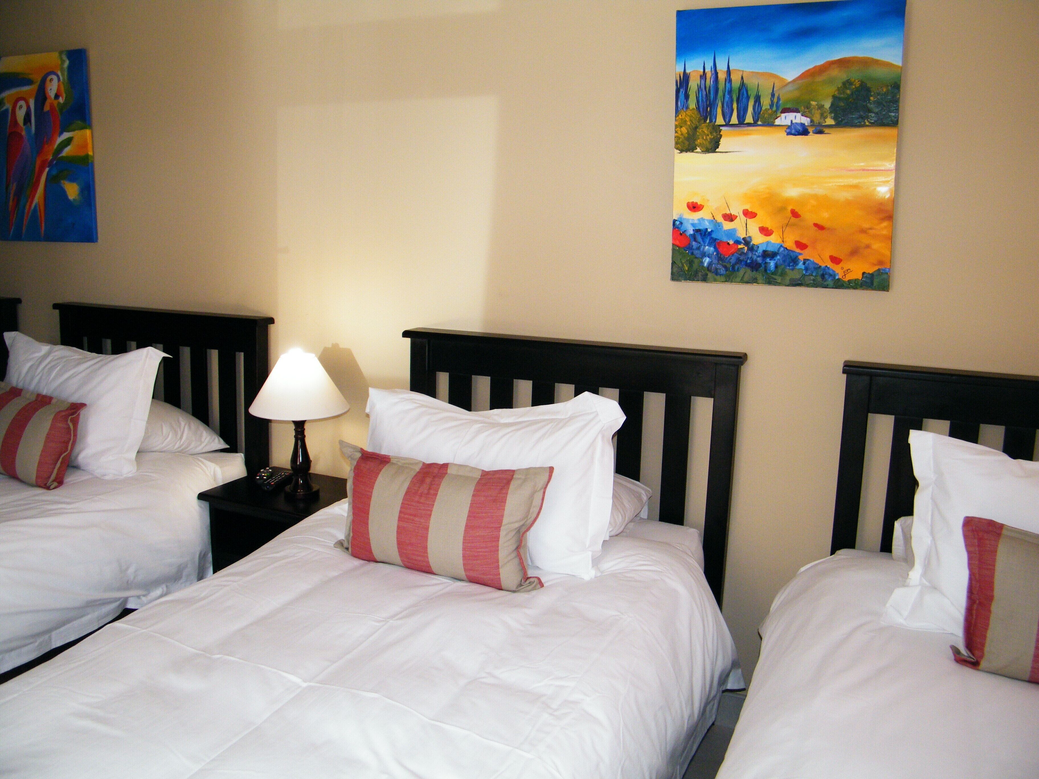 Habitación de hotel con tres camas individuales, cada una con ropa blanca y almohadas a rayas. Hay una mesita de noche con lámpara entre las camas.