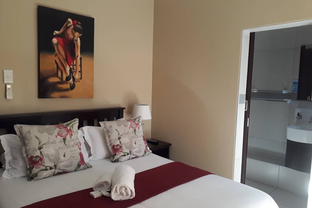 A hotel room with a bed, floral pillows, a red throw blanket, a painting on the wall, a bedside lamp, and a bathroom visible through an open door.