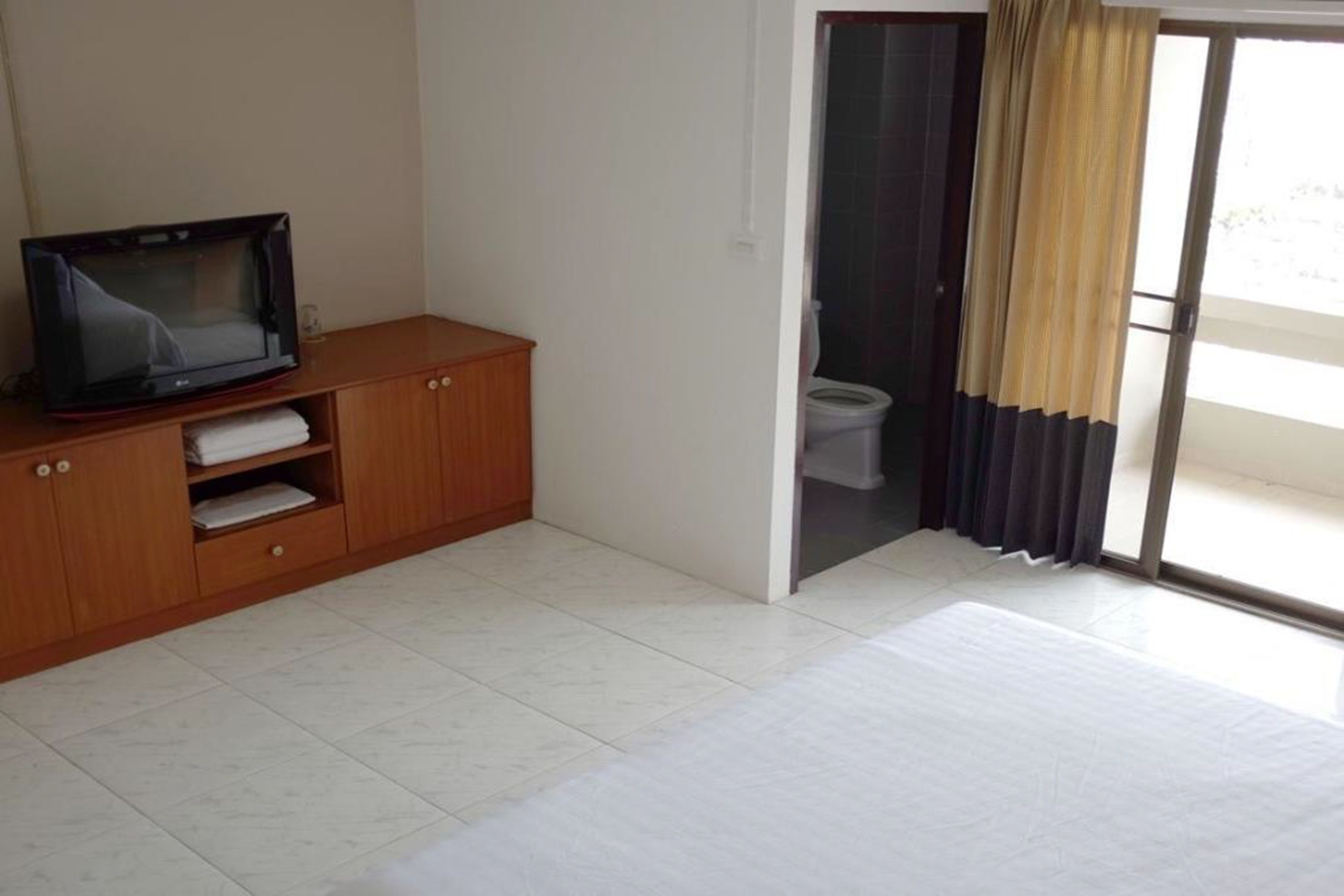 A bedroom with a wooden cabinet, a television, a sliding glass door leading to a bathroom, and a large window with curtains.