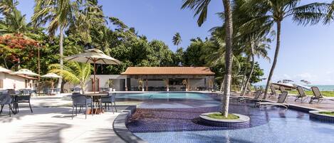 A pool area with lounge chairs and umbrellas, surrounded by palm trees and a building with a tiled roof.