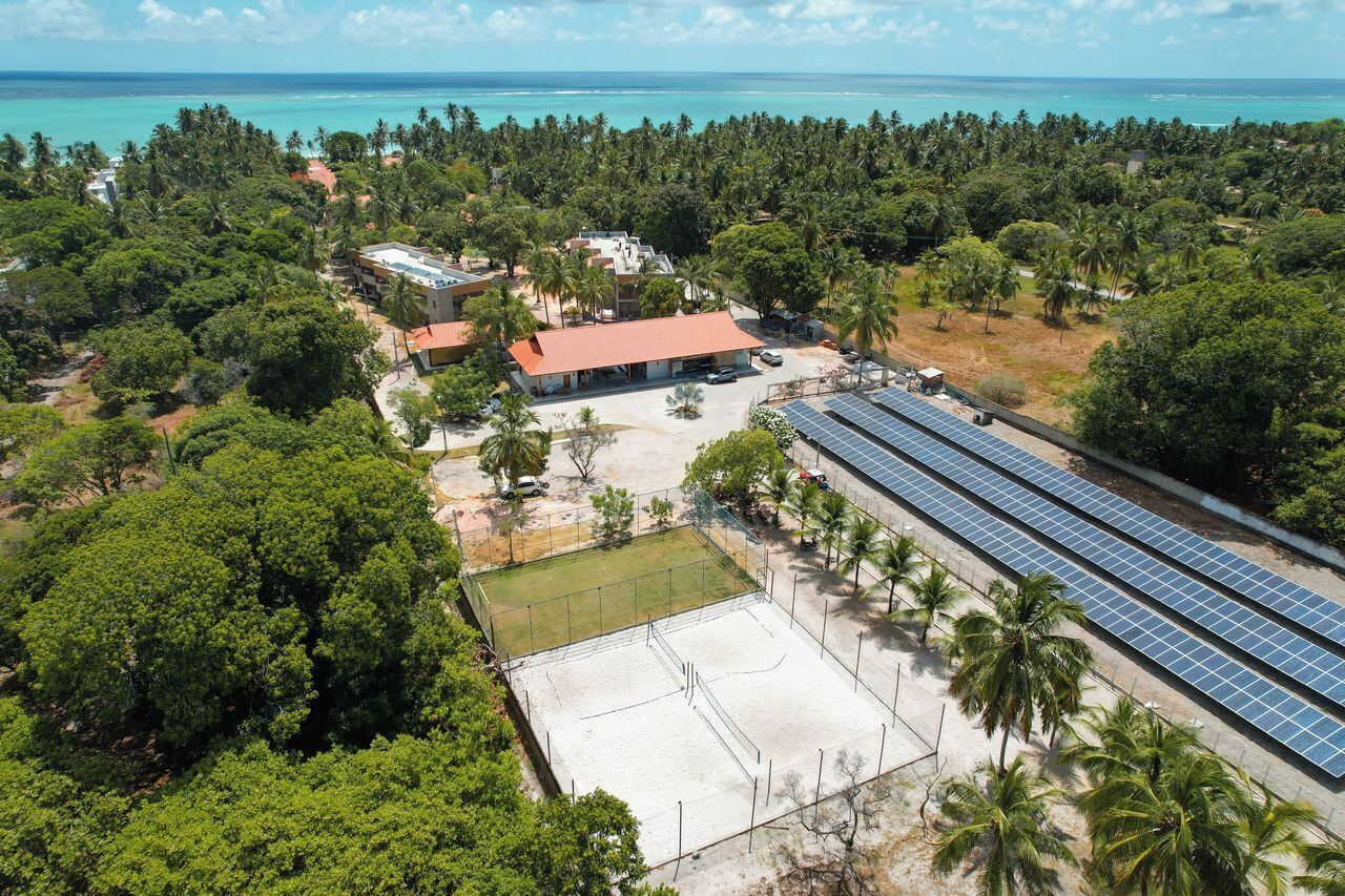 An aerial view of a resort with a large solar panel array, a tennis court, and a beachside location.