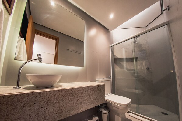 A modern bathroom with a granite countertop, a white sink, a mirror, a toilet, and a glass-enclosed shower. - Costa Dourada Village (Maragogi)