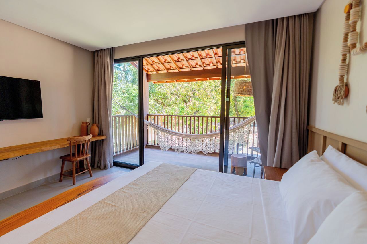 A hotel room with a large bed, a wooden chair, a small table, a TV mounted on the wall, and a balcony with a hammock.