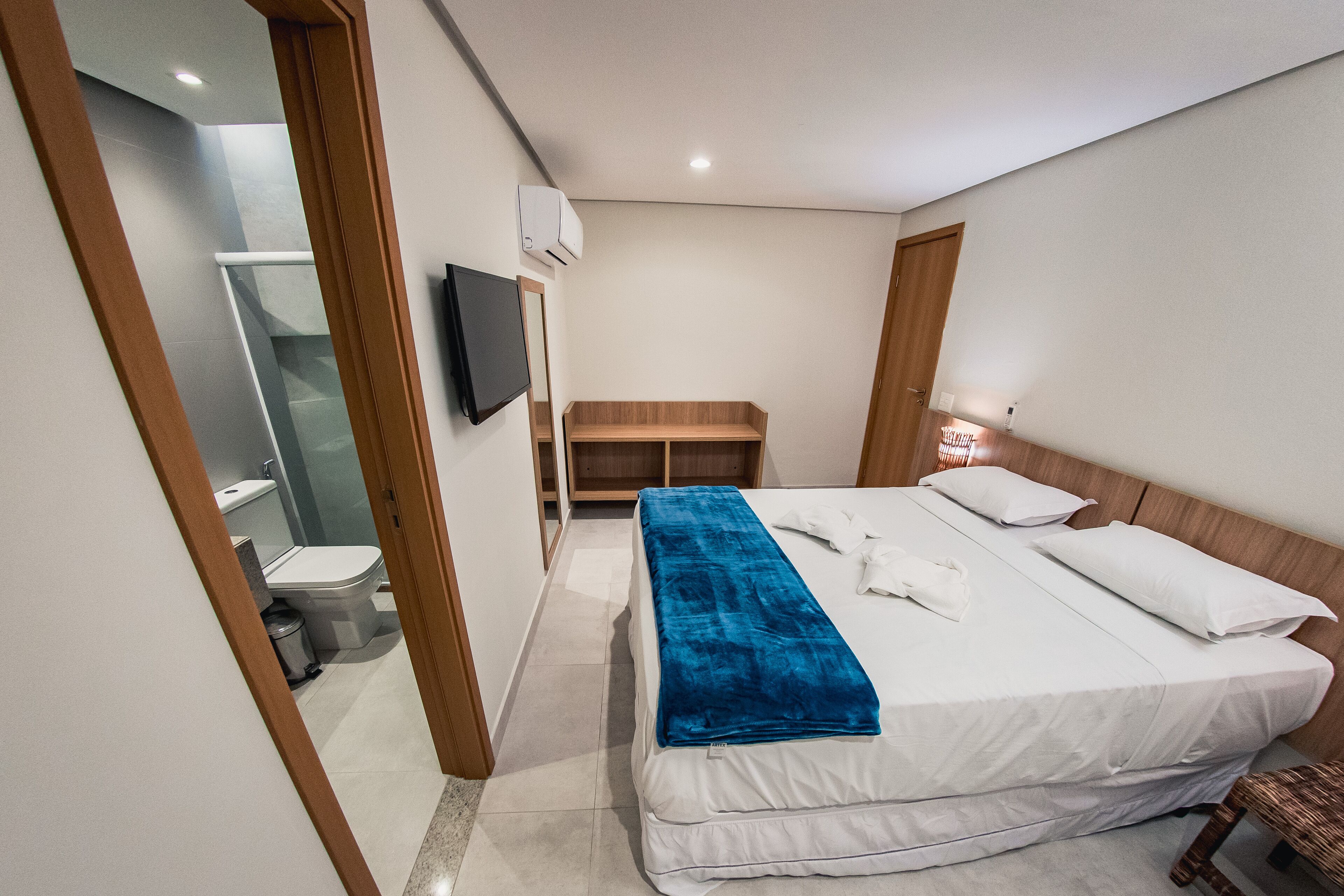 A hotel room with a bed, a TV, a wooden bench, and a bathroom visible through an open door.