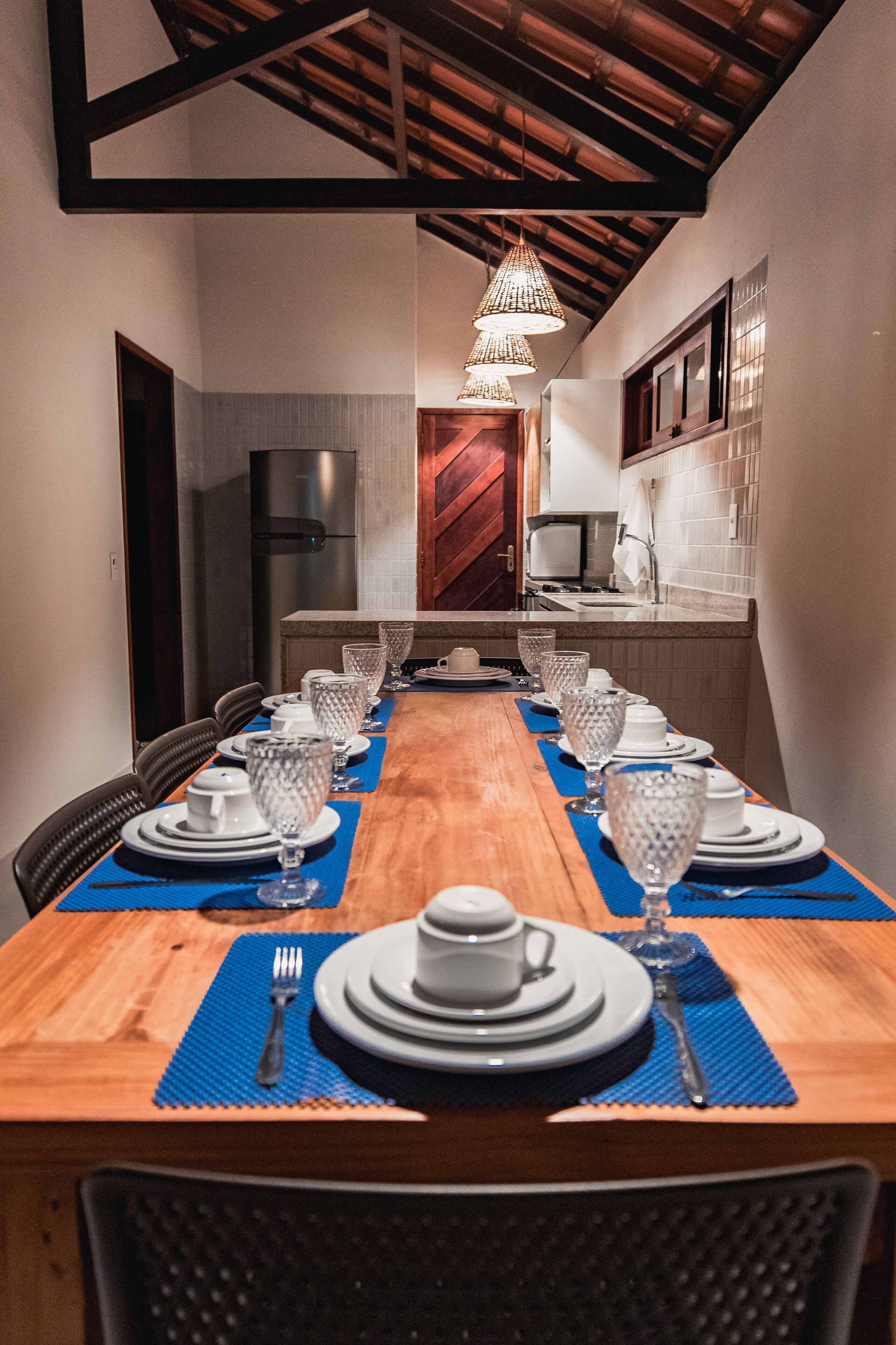 Un comedor con una larga mesa de madera dispuesta para una comida, con sillas a ambos lados, y una cocina al fondo.