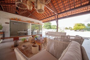 A modern outdoor lounge area with wicker furniture, a glass wall kitchen, and a view of the street. - Costa Dourada Village (Maragogi)